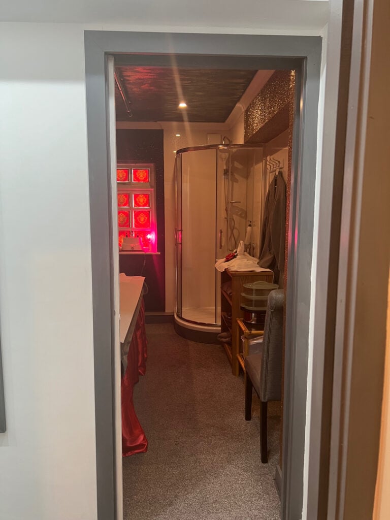 Cozy massage room with ambient lighting, featuring a massage table with a red cover, a shower stall, and a comfortable chair, creating a relaxing atmosphere for massage therapy in Woolmer Green.