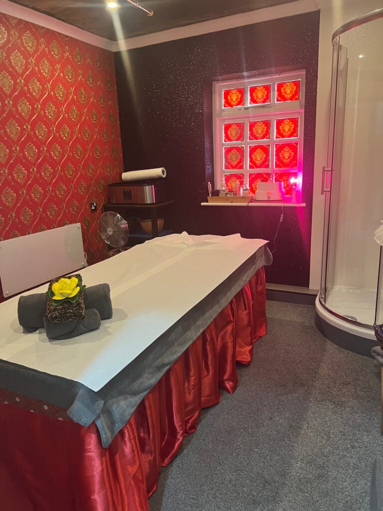 Massage therapy room with a massage table adorned with a yellow flower on grey towels, red curtains, soft lighting, and red-patterned wallpaper in Woolmer Green.