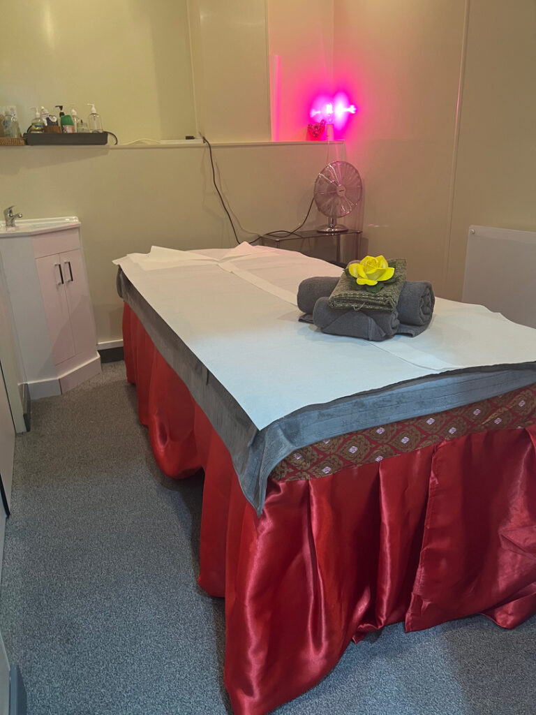 A cozy massage room in Woolmer Green with a neatly prepared massage table adorned with red silk draping, gray towels, and decorative elements, creating a tranquil ambiance under gentle lighting, perfect for therapeutic relaxation.