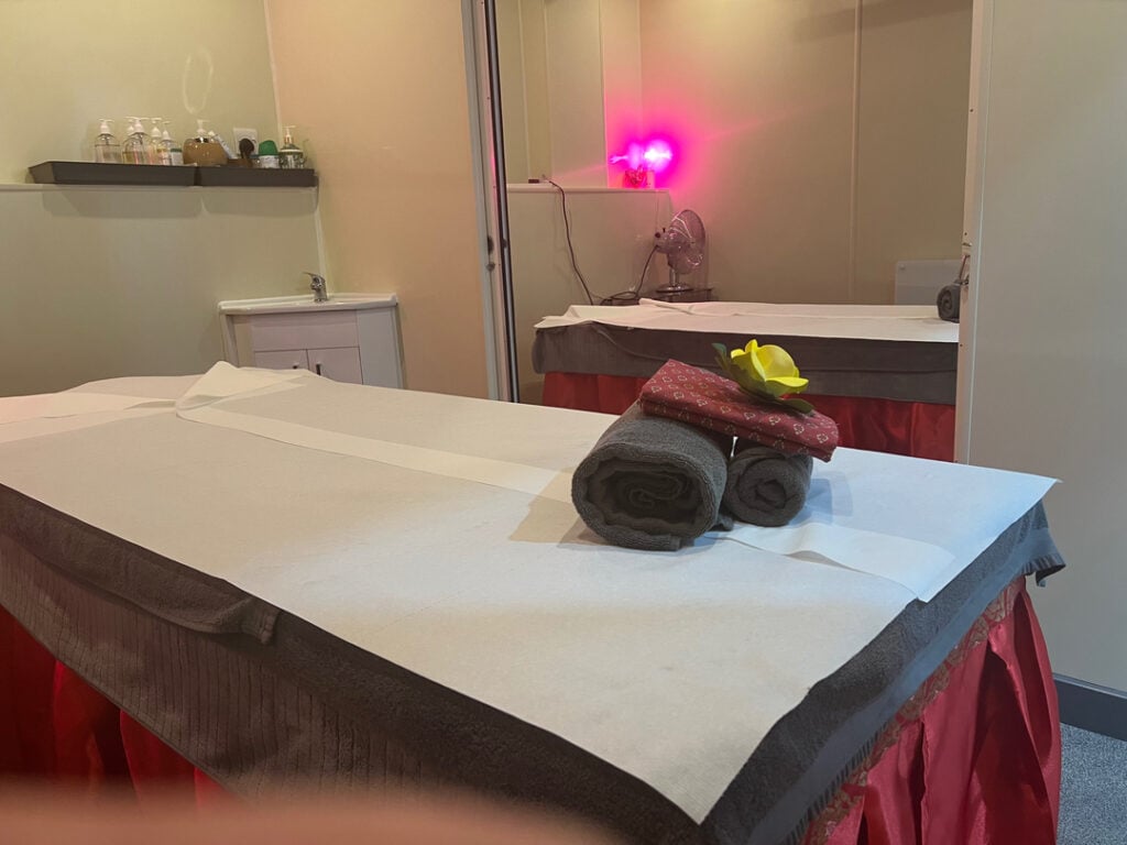 A serene massage spa room featuring neatly arranged towels with a decorative yellow flower atop a massage table, softly lit by a pink light, creating a calming ambiance perfect for relaxation and therapeutic massages in Knebworth.
