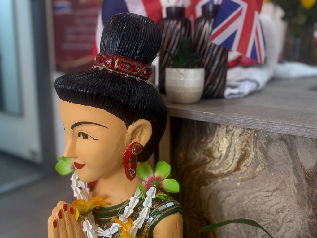 A traditional Thai statue adorned with floral garlands next to a counter displaying decorative items and Union Jack flags, representing a welcoming ambiance in a Woolmer Green massage spa.