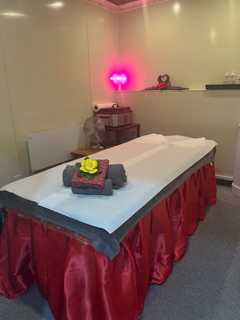 A tranquil massage therapy room in a peaceful setting with a neatly prepared massage table adorned with towels and a yellow flower, soft pink lighting, and a calming decorative touch, ideal for relaxation and rejuvenation in Knebworth.